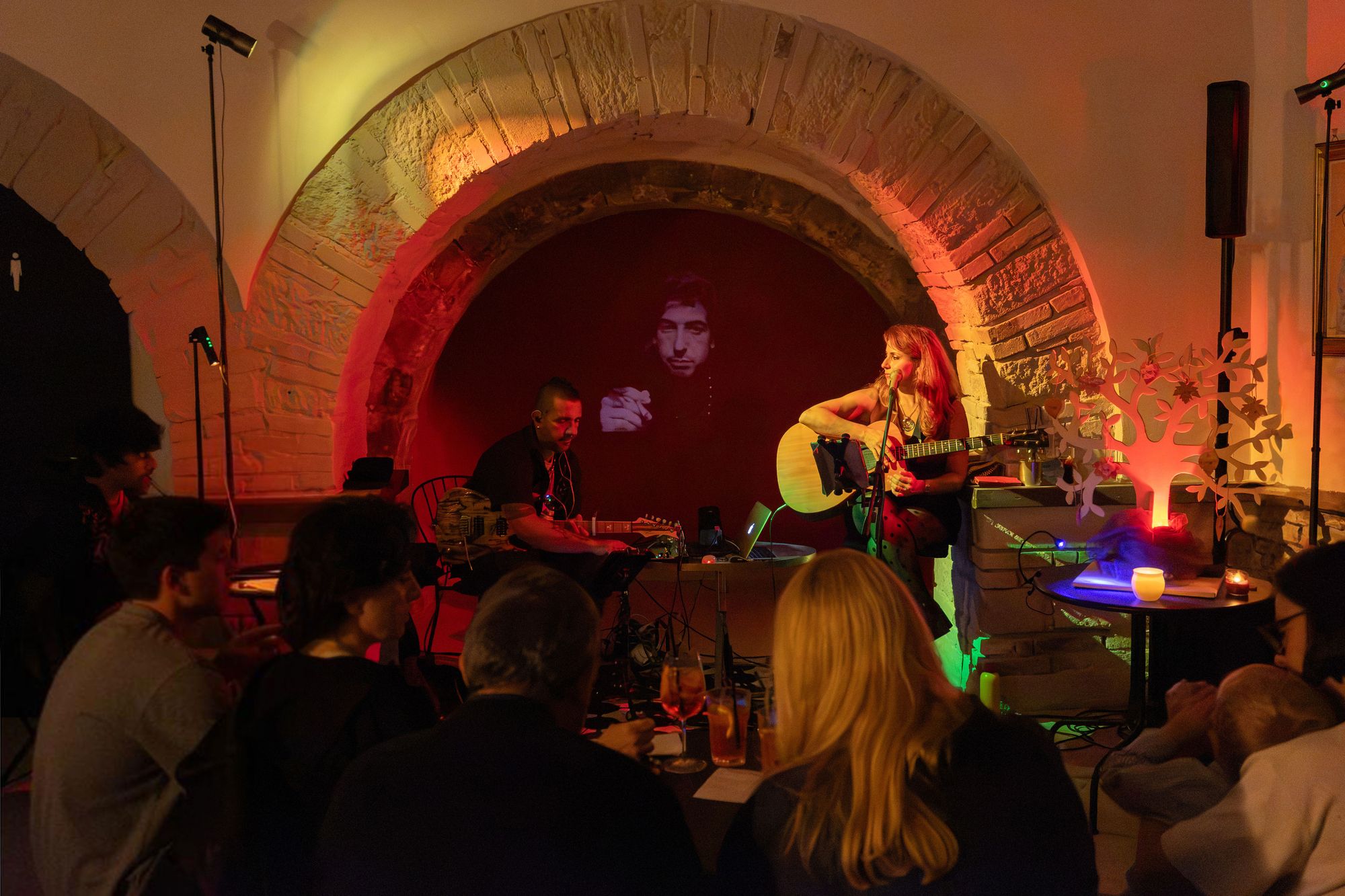 Lasimi canta alla chitarra sotto un arco di pietra, accanto all'albero di foglie; alle sue spalle è proiettato il volto di un artista